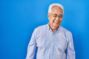 Hispanic senior man wearing glasses winking looking at the camera with sexy expression, cheerful and happy face. 