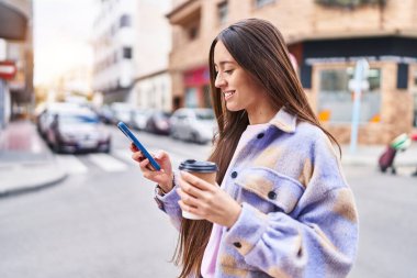Young beautiful hispanic woman using smartphone drinking coffee at street