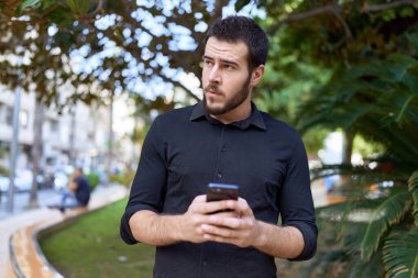 Genç İspanyol adam parkta rahat bir ifadeyle akıllı telefon kullanıyor.