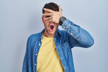 Young hispanic man standing over blue background peeking in shock covering face and eyes with hand, looking through fingers with embarrassed expression. 