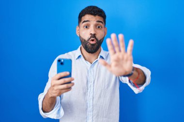 Hispanic man with beard using smartphone typing message doing stop gesture with hands palms, angry and frustration expression 