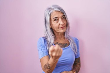 Middle age woman with tattoos standing over pink background beckoning come here gesture with hand inviting welcoming happy and smiling 