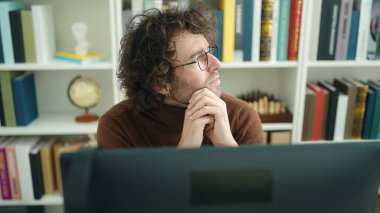 Young hispanic man student using computer with doubt expression at library university