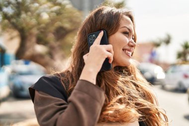 Genç, güzel, İspanyol bir kadın gülümsüyor ve caddede akıllı telefondan konuşuyor.