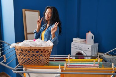 Young asian woman hanging clothes at clothesline smiling looking to the camera showing fingers doing victory sign. number two. 