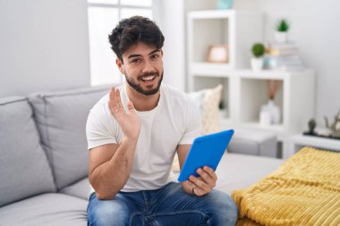 Hispanic man with beard using touchpad sitting on the sofa waiving saying hello happy and smiling, friendly welcome gesture 
