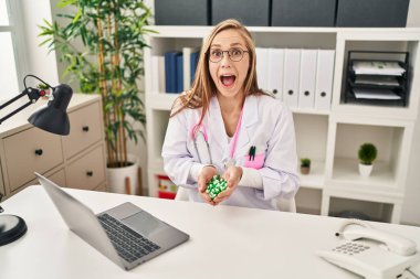 Young blonde doctor woman holding pills celebrating crazy and amazed for success with open eyes screaming excited. 