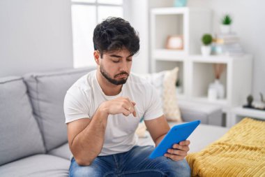 Hispanic man with beard using touchpad sitting on the sofa pointing down looking sad and upset, indicating direction with fingers, unhappy and depressed. 