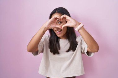 Young hispanic woman standing over pink background doing heart shape with hand and fingers smiling looking through sign 