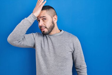 Hispanic man standing over blue background surprised with hand on head for mistake, remember error. forgot, bad memory concept. 