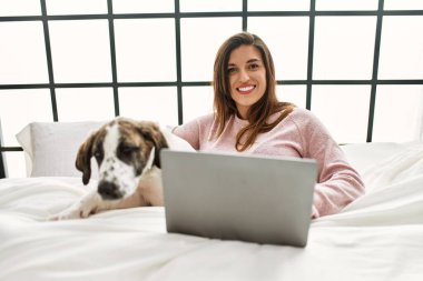 Yatak odasında dizüstü bilgisayar kullanan, köpeğini kucaklayan genç bir kadın.