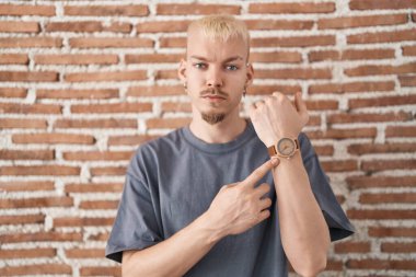 Young caucasian man standing over bricks wall in hurry pointing to watch time, impatience, looking at the camera with relaxed expression 