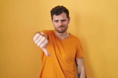 Young hispanic man standing over yellow background looking unhappy and angry showing rejection and negative with thumbs down gesture. bad expression. 