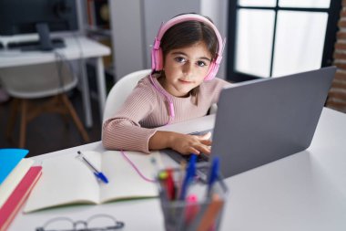 Adorable hispanic girl student using laptop and headphones sitting on table at classroom