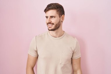 Hispanic man with beard standing over pink background looking away to side with smile on face, natural expression. laughing confident. 