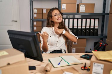 Young hispanic woman working at small business ecommerce pointing to the back behind with hand and thumbs up, smiling confident 