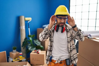 Young hispanic man with beard working at home renovation trying to hear both hands on ear gesture, curious for gossip. hearing problem, deaf 