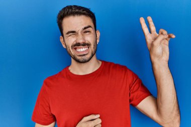 Young hispanic man with beard wearing red t shirt over blue background smiling with happy face winking at the camera doing victory sign. number two. 