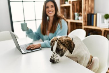 Evde köpekle aynı masada oturan dizüstü bilgisayar kullanan genç bir kadın.