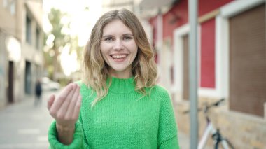 Genç sarışın kadın gülümsüyor. Kendinden emin. Sokakta el kol hareketi yapıyor.