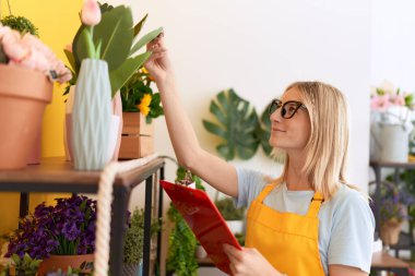 Young blonde woman florist reading document touching plant at flower shop