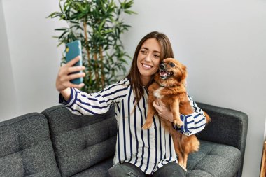 Genç İspanyol kadın akıllı telefonun başında selfie çekiyor. Kanepede oturup evdeki köpeğe sarılıyor.