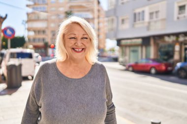 Orta yaşlı sarışın kadın sokakta kendinden emin bir şekilde gülümsüyor.