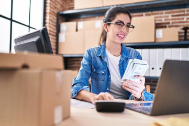 Young beautiful hispanic woman ecommerce business worker holding dollars using calculator at office