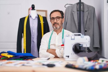 Middle age man tailor sitting on table with serious expression and arms crossed gesture at tailor shop