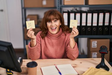 Middle age hispanic woman working at small business ecommerce with happy and sad emotion smiling and laughing hard out loud because funny crazy joke. 