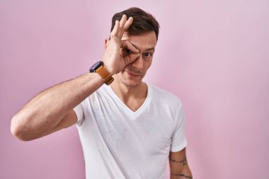 Caucasian man standing over pink background doing ok gesture with hand smiling, eye looking through fingers with happy face. 