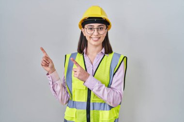 Hispanic girl wearing builder uniform and hardhat smiling and looking at the camera pointing with two hands and fingers to the side. 