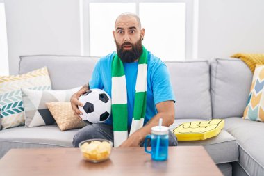 Young hispanic man with beard and tattoos football hooligan holding ball supporting team scared and amazed with open mouth for surprise, disbelief face 