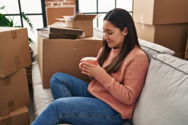 Young hispanic woman drinking coffee sitting on sofa at new home