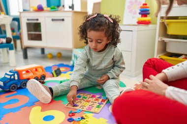Adorable hispanic girl playing with maths puzzle game sitting on floor at kindergarten