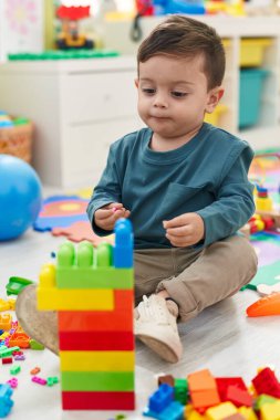 Adorable hispanic boy playing with construction blocks sitting on floor at kindergarten