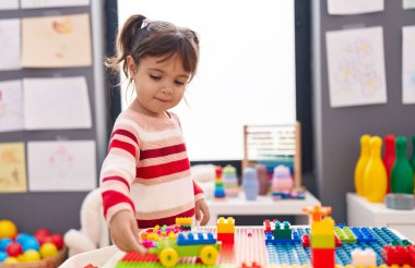 Adorable hispanic girl playing with construction blocks standing at kindergarten