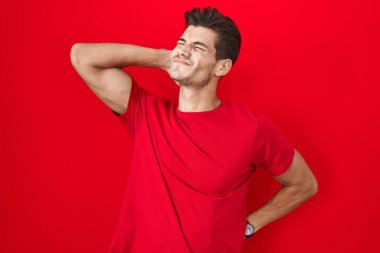 Young hispanic man standing over red background suffering of neck ache injury, touching neck with hand, muscular pain 
