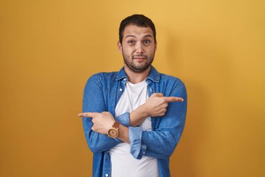 Hispanic man standing over yellow background pointing to both sides with fingers, different direction disagree 