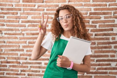 Young caucasian woman holding art notebook smiling with happy face winking at the camera doing victory sign with fingers. number two. 