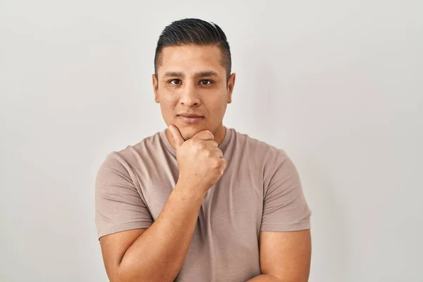 Hispanic young man standing over white background looking confident at the camera with smile with crossed arms and hand raised on chin. thinking positive.