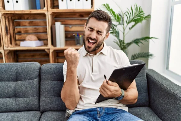 Handsome hispanic man holding clipboard working at psychology clinic very happy and excited doing winner gesture with arms raised, smiling and screaming for success. celebration concept.