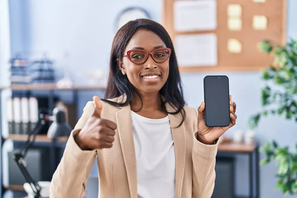 African young woman holding smartphone showing blank screen smiling happy and positive, thumb up doing excellent and approval sign