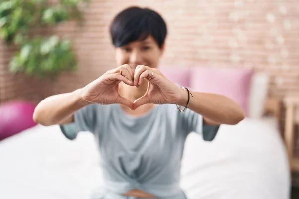 Middle age chinese woman doing heart gesture sitting on bed at bedroom