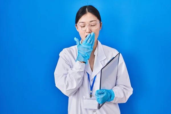 Chinese young woman working at scientist laboratory bored yawning tired covering mouth with hand. restless and sleepiness.