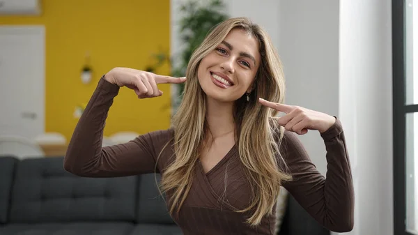 Young blonde woman pointing to mouth with fingers sitting on sofa at home