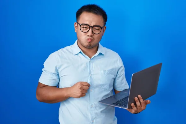 Chinese young man using computer laptop puffing cheeks with funny face. mouth inflated with air, crazy expression.