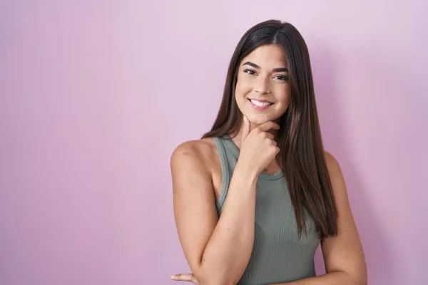 Hispanic woman standing over pink background looking confident at the camera smiling with crossed arms and hand raised on chin. thinking positive.
