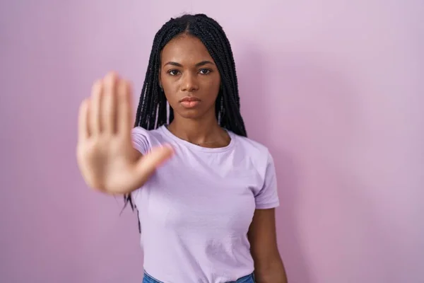 African american woman with braids standing over pink background doing stop sing with palm of the hand. warning expression with negative and serious gesture on the face.