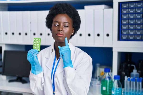 African young woman working at scientist laboratory holding birth control pills serious face thinking about question with hand on chin, thoughtful about confusing idea